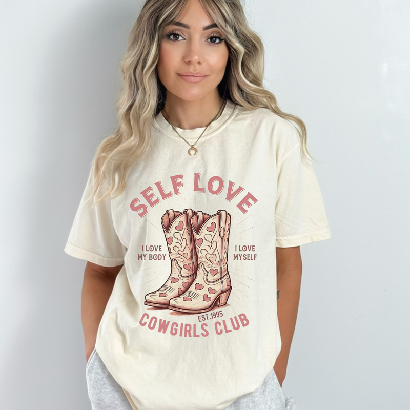 Cream oversized vintage-style t-shirt featuring a graphic of cowgirl boots with pink hearts and the phrase “Self Love Cowgirl Club – I love my body, I love myself.” Western-inspired design with a bold, empowering message.
